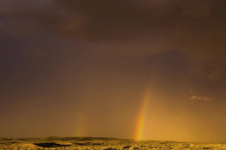 Two rainbows stand together in a dark yellow surreal sky as clouds move overhead and a light glows along the horizon