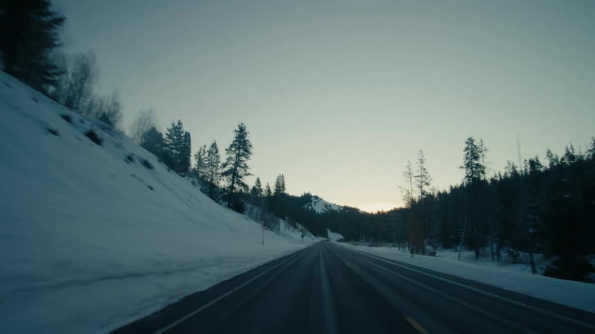 Vehicle drives along country road near snowy slope and green forest at dusk