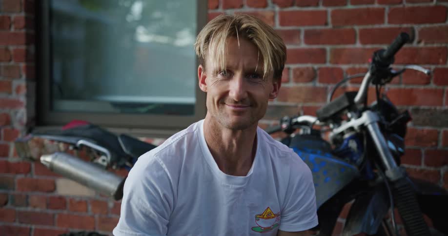 Blonde man sits outside building near motorcycle smiling