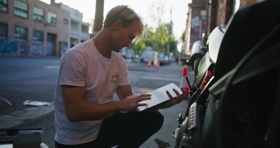 A man is holding a clipboard and writing as he kneels down looking at a motorcycle outside of a building