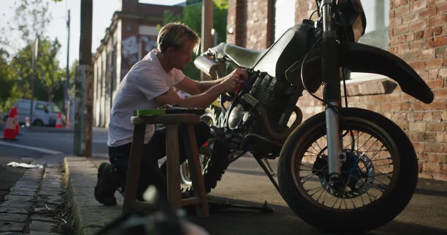 Adult male with blond hair squatting on the sidewalk while working on his motorcycle with tools