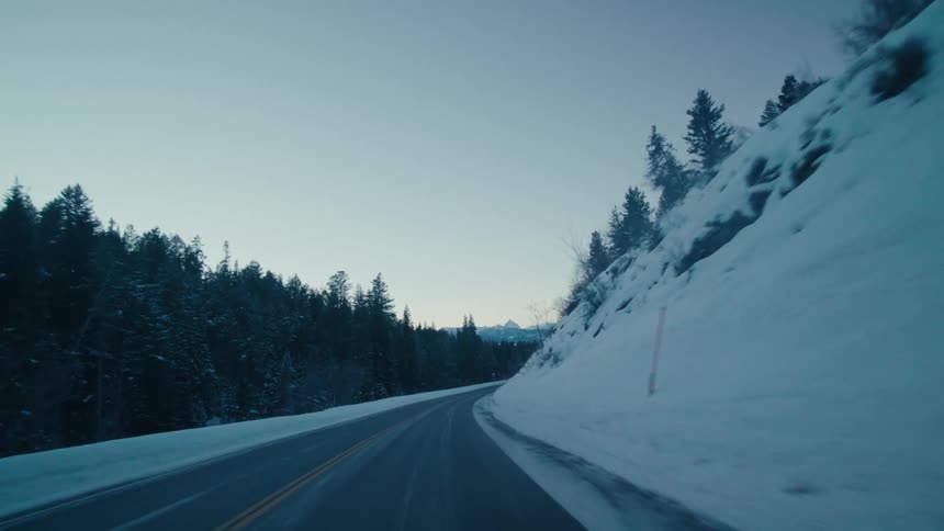 Road passes between conifer forest and snow covered slope on route to mountains on the horizon