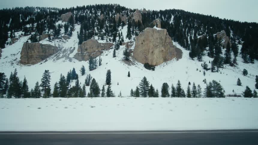The camera is moving right like someone is driving down a street looking at snowcovered mountains and pine trees