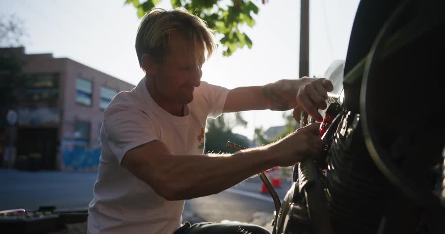 A man is kneeling down on the side of the road working on a motorcycle and he puts a wrench back in his tool box