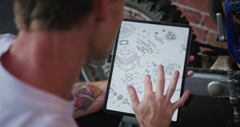 A man working on his motorcycle reviews a manual on his tablet