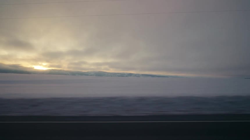 Sun sets as vehicles move along highway past snow covered fields near silos