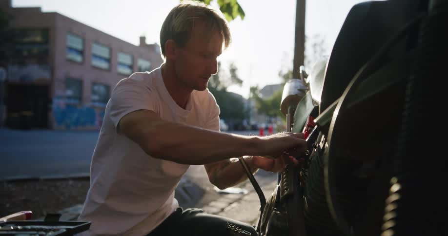 Blonde man with white t shirt and black pants fixes motorcycle in the street