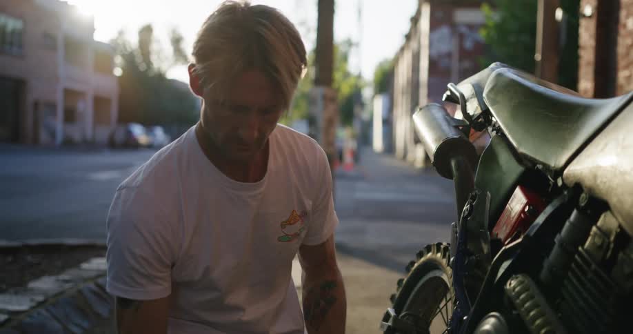 Blonde man with tattoos and white t shirt fixes bike near building in the street