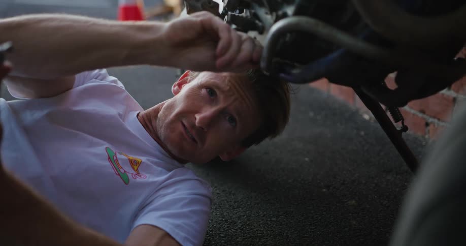 Blonde man with white t shirt and tattoos lies on the ground fixing motorcycle