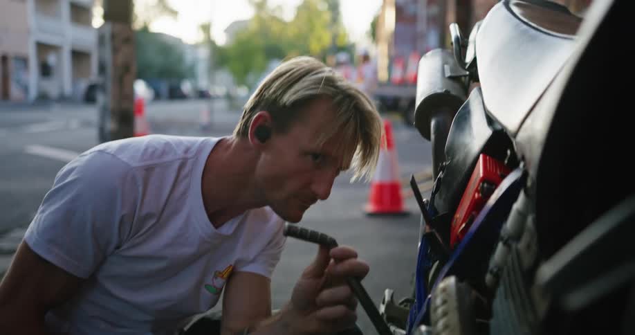 Blonde man with white t shirt and tattoos fixes motorcycle in the street