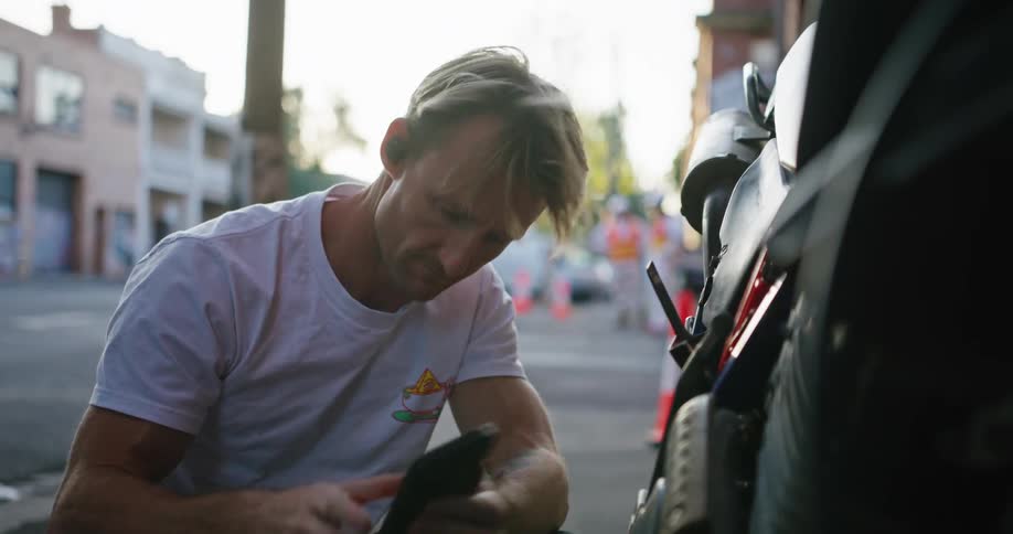 Man works listens to music while working on motorcycle as road construction workers stand on opposite corner