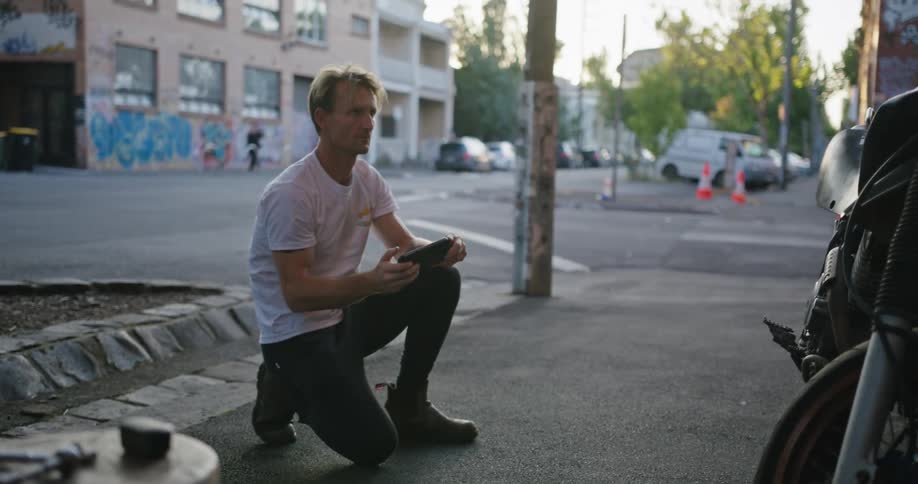 Blonde man with white t shirt an jeans photographs motorcycle with smartphone in the street