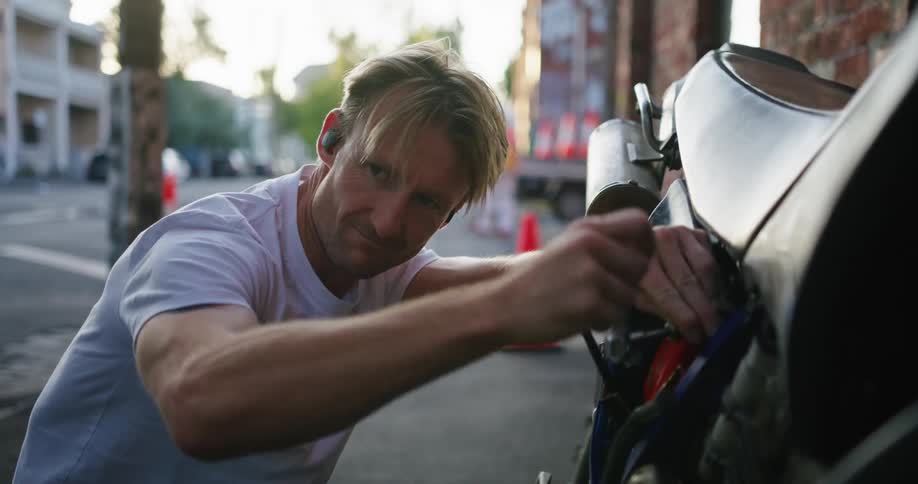 Blonde man with white shirt and tattoos fixes motorcycle in the street