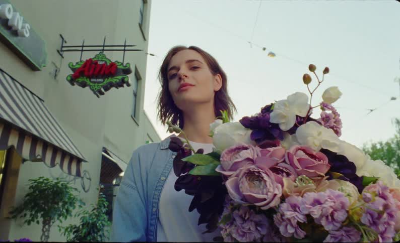 Woman with brown hair denim shirt and white top holds a bunch of flowers and smiles at camera near the flower store