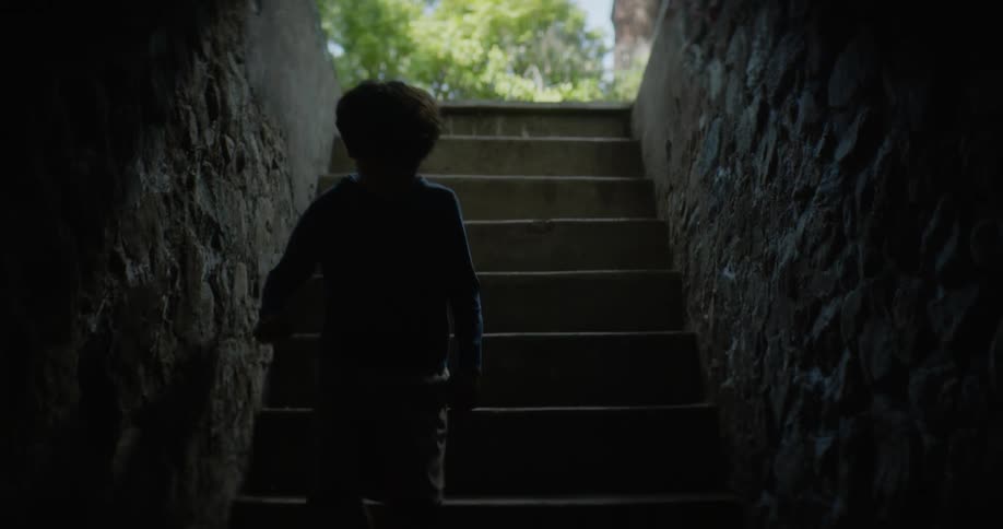 A little boy wearing big tennis shoes walks down a dark stairway leading under a building and he stops at the bottom