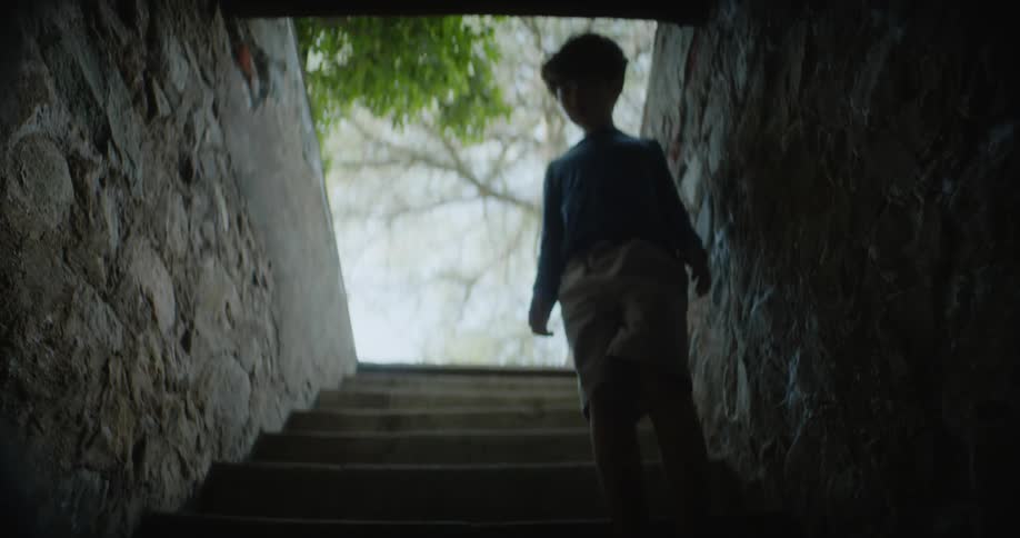 A young boy wearing big tennis shoes walks down a dark outdoor stairway and he stops to reach his hand out