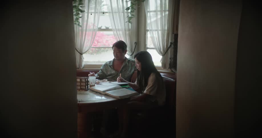 Man in blue shirt sits at table with daughter with long straight hair writing in notebook