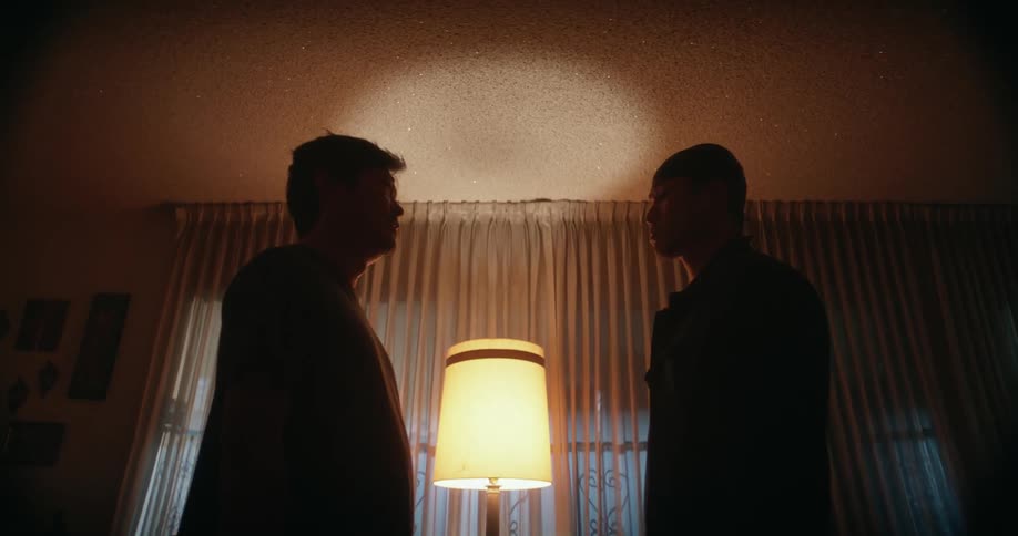 Two men standing in living room in sillhouette nervously talking about something as headlights shine through window