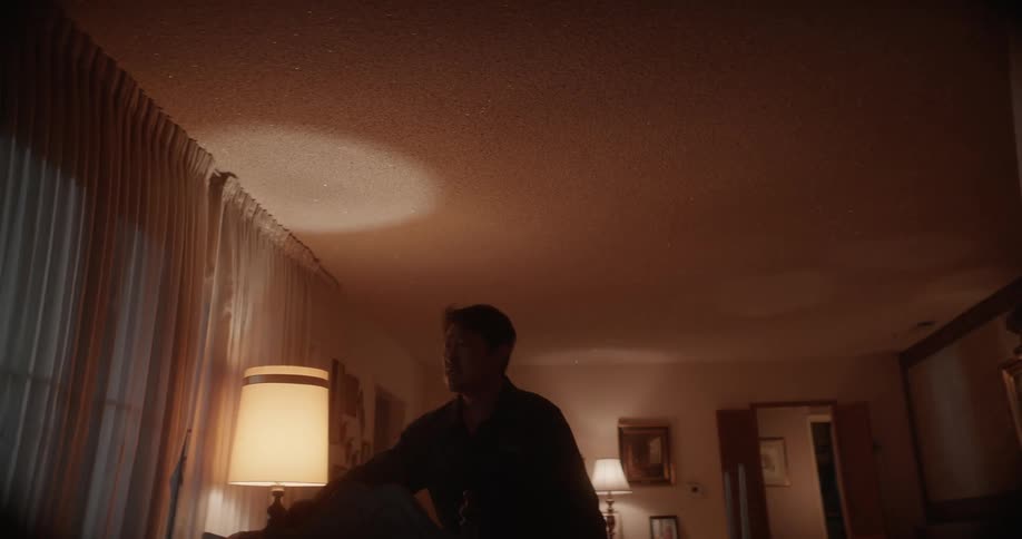 A man walks through his living room to the window as he pulls back the curtain and looks outside