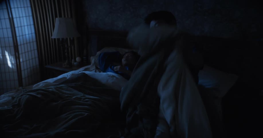 Man sitting on side of bed in dark bedroom gets into bed with sleeping wife