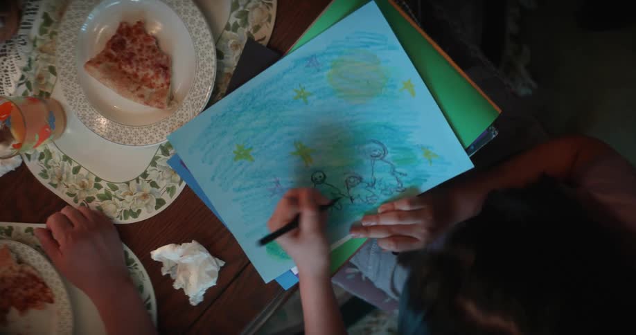 Girl sits at table drawingnext to plate holding a partially eaten slice of pizza