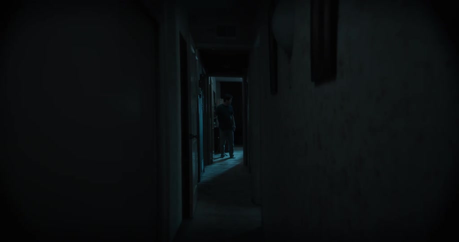 A man stands in the doorway of a room and walks down the dark hallway