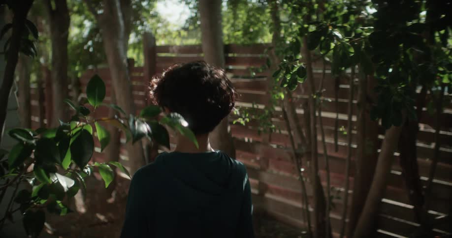 Boy wearing a hoodie walks through a backyard between a building and a fence