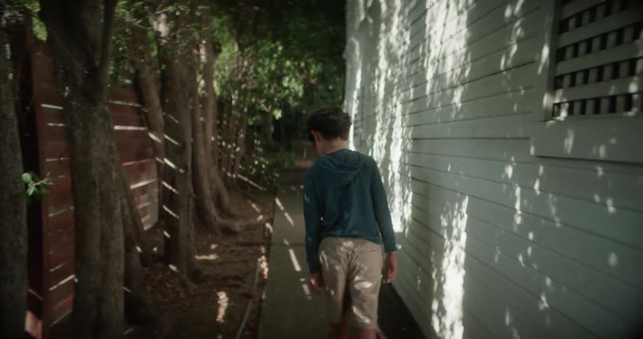 Boy walks along the side of a house and a fence in the tree lined yard