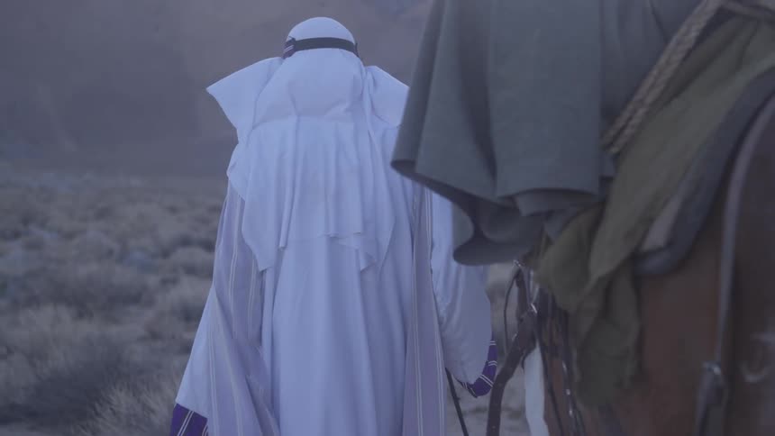 A man in a white robe leads a horse through the desert