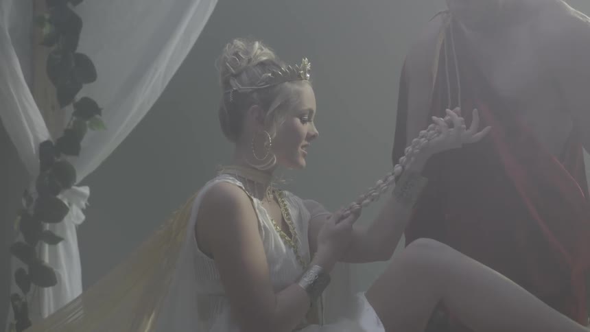 Goddess lounging on a bed while someone approaches and gives her a necklace