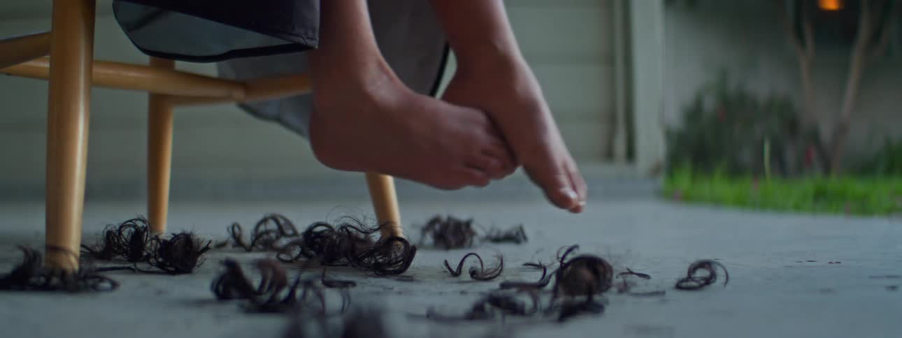 A childs bare feel are moving as they sit on a stool outside in their driveway getting a hair cut as locks of hair fall to the ground