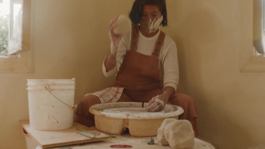 A woman sitting in the corner of a room grabs a piece of clay and starts working it on a pottery wheel