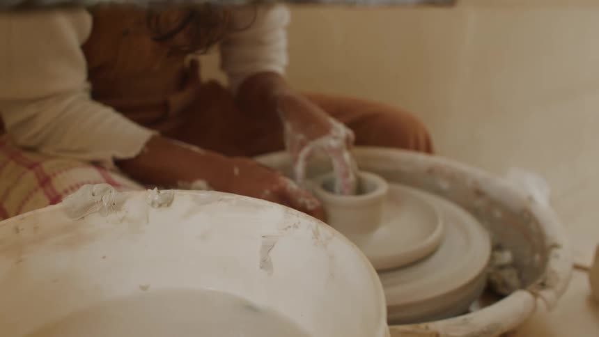 A potter is sitting at a pottery wheel working with wet clay as they dip their hand into a bowl of water