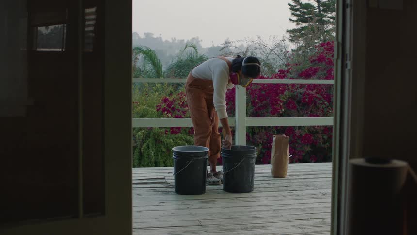 A woman with a mask on stands on her balcony and stirs something in a big pail