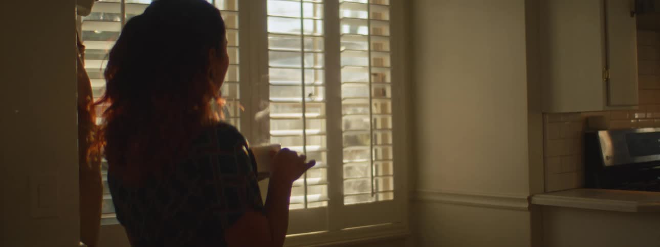 A woman stands next to her kitchen window holding a hot cup of coffee