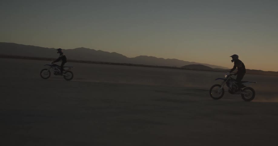Two cyclists are riding motor bikes across the desert as the sun shines brightly on the horizon