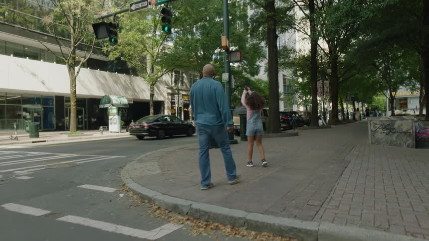 A father runs across a city street with her daughter as she holds up a cell phone and takes a picture