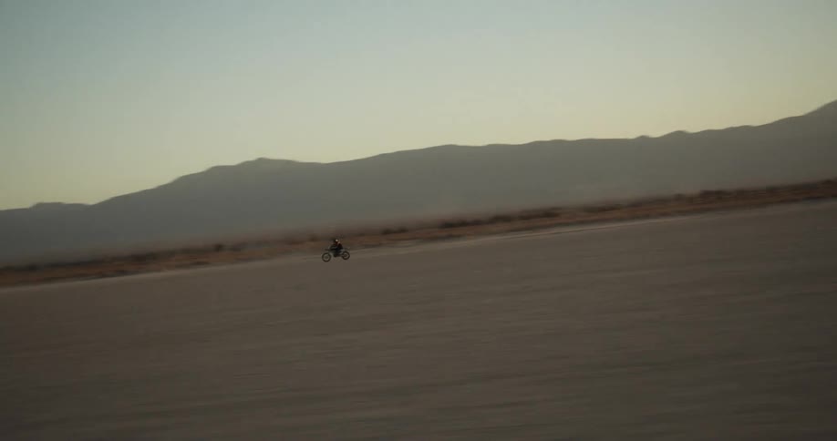 Person on a motorcycle riding through the desert at dusk is filmed from a vehicle moving in the opposite direction