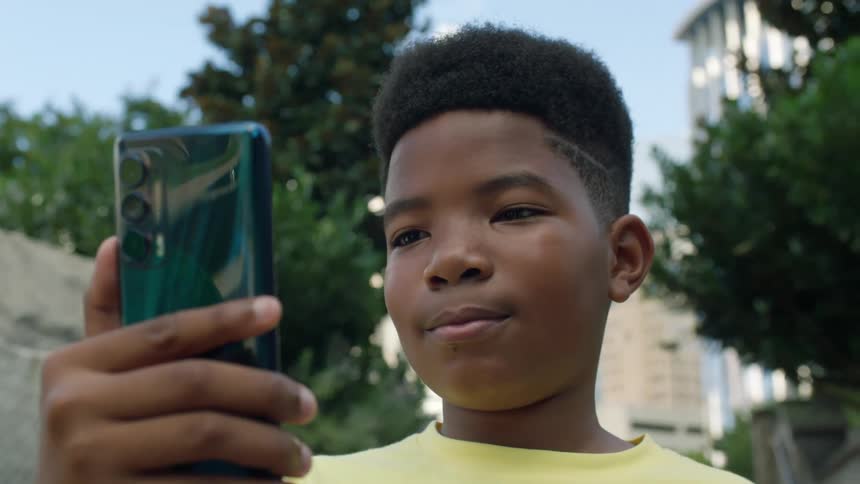 A young boy stands outside and looks at a cell phone