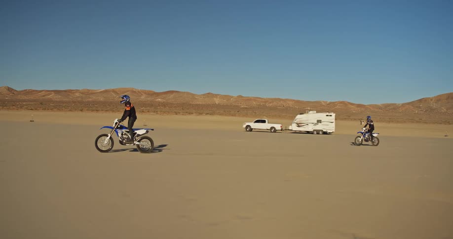 Two men on motorbikes ride past a camper in the desert