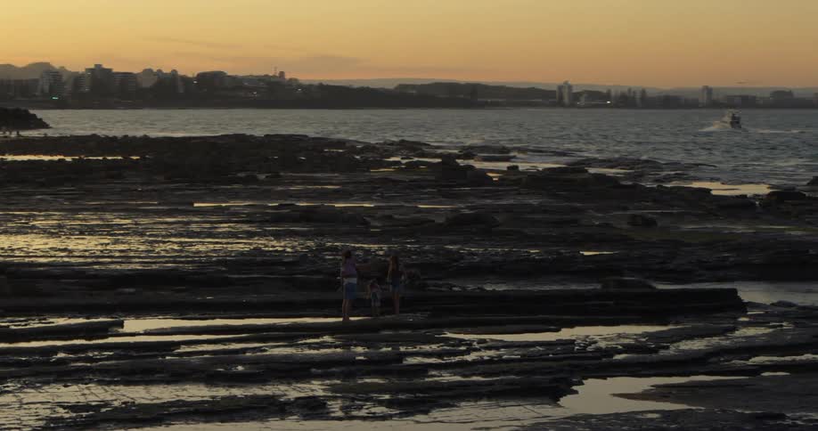 Family stands on beach near tidal pools as boat leaving wake moves behind them toward city skyline
