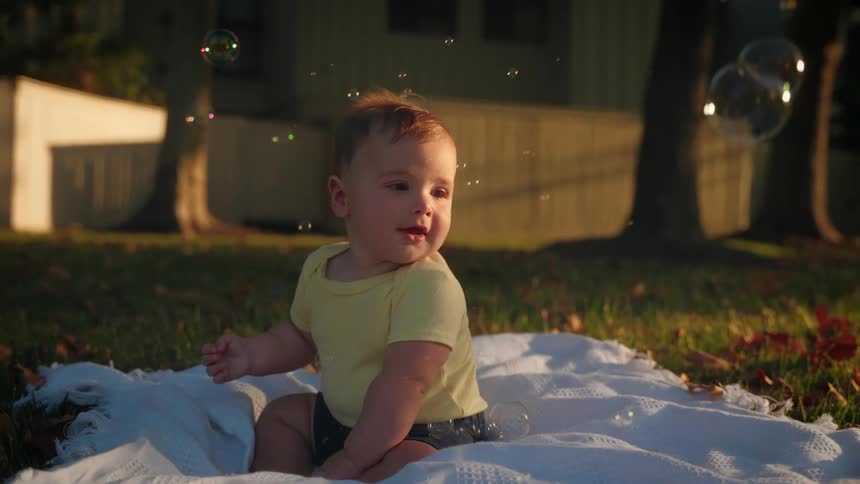 A baby sits on a blanket in his yard and smiles as bubbles float around him