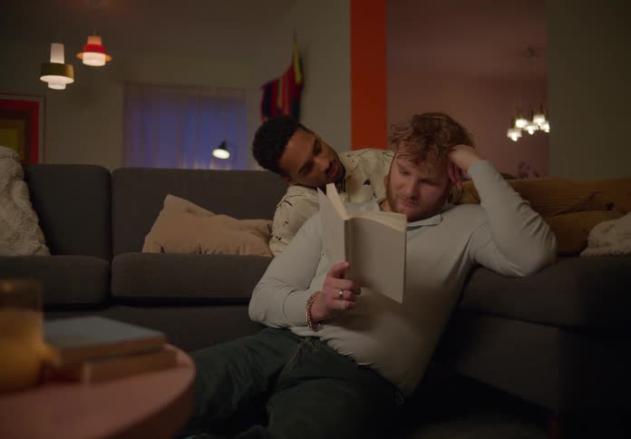 One man reads while another man touches his shoulder and kisses him then they exchange smiles