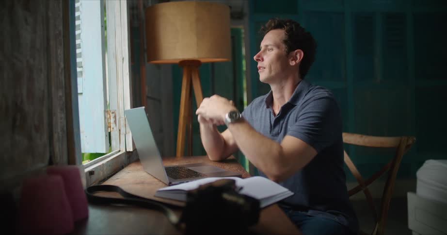 A man is sitting at a desk in front of a window looking at an open book as he types on a laptop computer