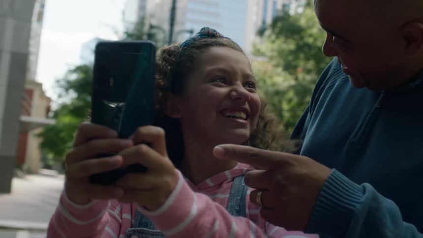A young girl is standing on a downtown street with her father as she holds up her phone and he points to the screen