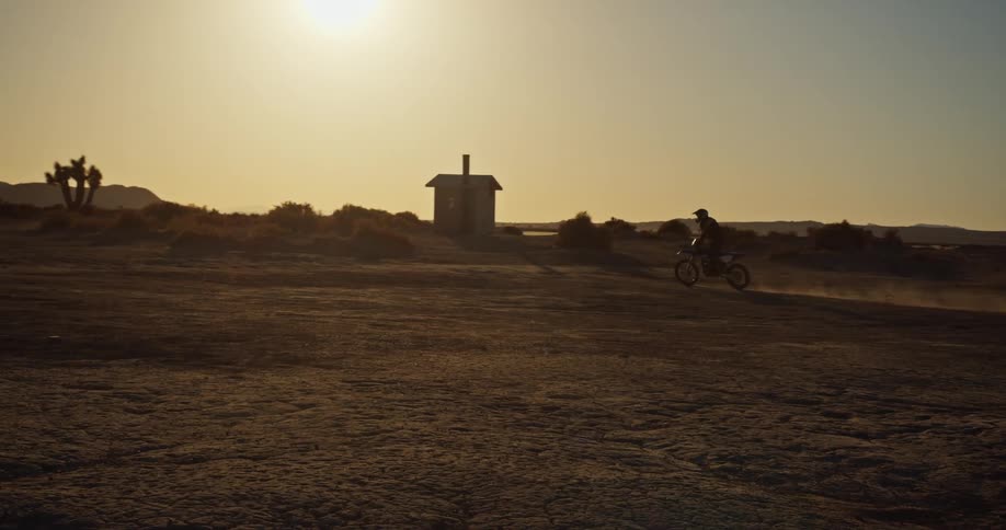 A cyclist is riding a motor bike across the desert and there is a small building and mountains in the distance