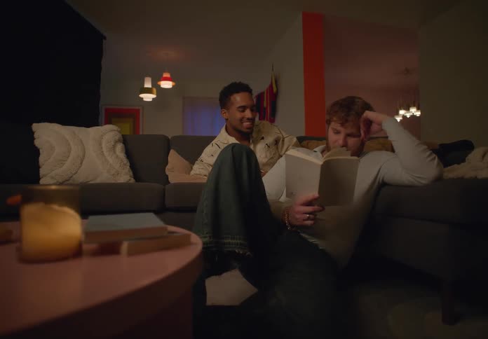 A man reading on the floor while another man jumps on the couch behind him