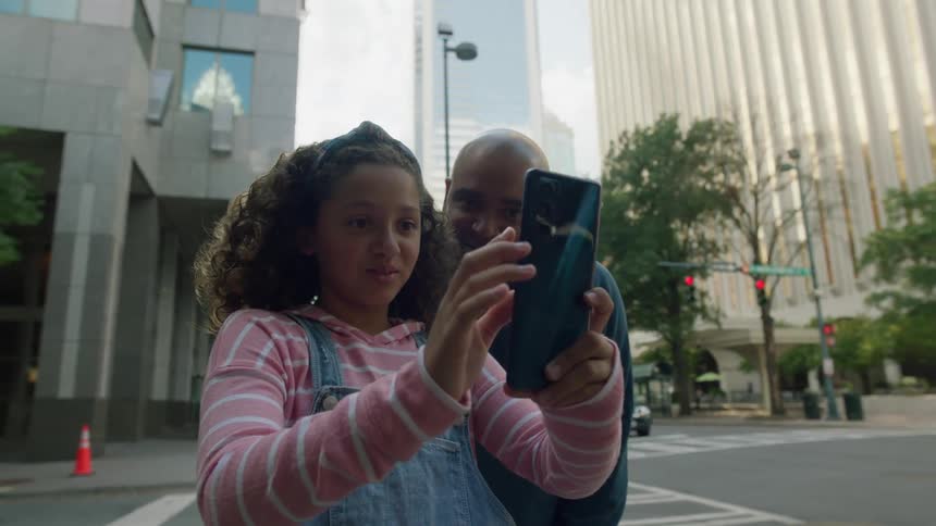 Father leans over to talk to his daughter and share her smartphone as they stand on downtown corner