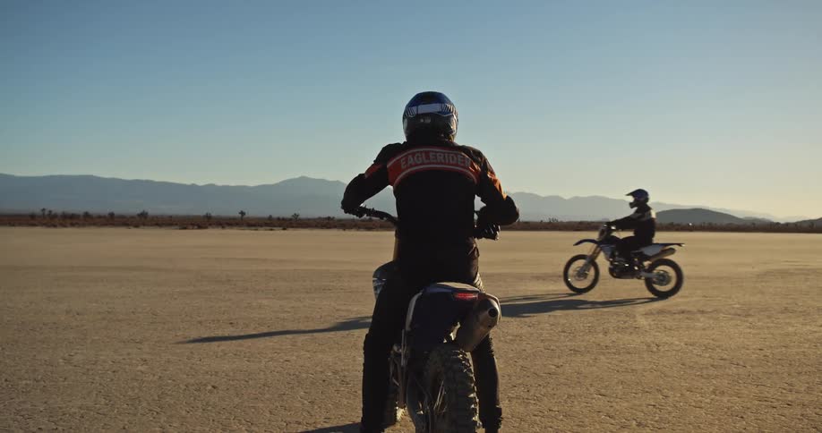 Two people on motorcycles slowly drive in a desert area