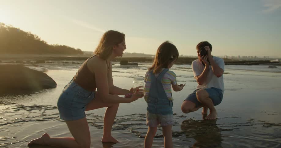 Father videotapes as mother holds jar for daughter to put findings she picks up from beach into with skyline on misty horizon